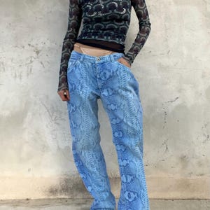Op de afbeelding: Een persoon draagt een top met lange mouwen met patroon en een lichtblauwe jeans met slangenprint. De top heeft een hoge hals en een doorschijnend, patroon. De jeans heeft een losse pasvorm met een slangenpatroon.