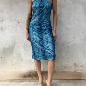 Op de afbeelding: Een knielange mouwloze jurk met een levendig blauw en zwart slangenprintpatroon. Het kledingstuk heeft een hoge halterhalslijn met een kleine druppelvormige uitsnede in het midden van de borst, wat zorgt voor een nauwsluitend silhouet.