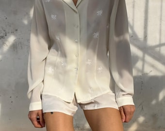 Könnte beinhalten: Ein cremefarbenes, langärmeliges Hemd mit Knopfleiste und zartem, weißem Blumenstickmuster, kombiniert mit passenden cremefarbenen Shorts. Das Hemd hat einen klassischen Kragen und eine durchgehende Knopfleiste mit durchscheinenden Knöpfen.
