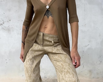 Könnte beinhalten: Eine braune, gerippte Strickjacke mit Dreiviertelärmeln und einem einzelnen weißen Knopfverschluss an der Brust, kombiniert mit einer beigen Hose mit tonalen Damast-Blumenmuster und einem strukturierten Bund mit Gürtelschlaufen.