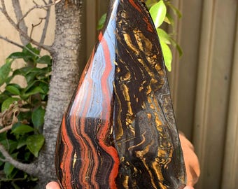 May include: A polished, teardrop-shaped stone with a striking pattern of red, black, gold, and blue bands. The stone's surface is smooth and reflective, showcasing the natural colors and textures of the mineral. The stone is held in a hand.