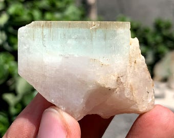 May include: A raw, natural aquamarine crystal with a pale blue-green hue. The crystal has a rectangular shape with a rough, textured surface and a milky white base. The top portion of the crystal is a translucent, light blue-green color.