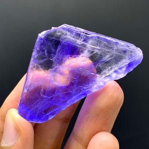 May include: A translucent, crystalline mineral specimen with a vibrant purple hue. The stone has a rough, irregular shape and appears to be held in a hand against a dark background. The color is concentrated in the center, fading towards the edges.
