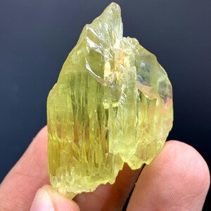 Könnte beinhalten: Eine rohe, natürliche gelb-grüne Kristallformation. Das Mineral hat eine raue, strukturierte Oberfläche mit sichtbaren kristallinen Strukturen. Die Farbe ist ein leuchtendes, durchscheinendes Gelbgrün, wobei einige Bereiche undurchsichtiger erscheinen. Der Kristall wird in einer Hand gehalten.