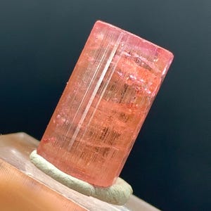 Pode incluir: Um cristal rosa translúcido e retangular com estrias verticais. O cristal é montado em uma base de cor clara com um adesivo cinza. O fundo é de cor escura e sólida.