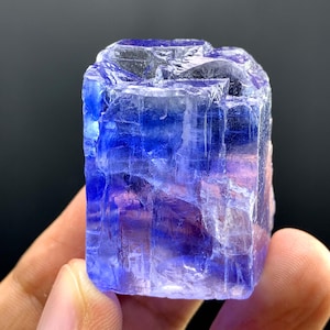 Puede incluir: Un vibrante cristal de tanzanita azul-violeta. La gema tiene forma rectangular con facetas visibles y un aspecto translúcido. El color transita del azul profundo a tonos más claros, con toques de púrpura y rosa. El cristal se sostiene en una mano.