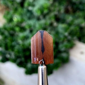 Puede incluir: Un pequeño trozo rectangular de madera con una franja oscura vertical en el centro. La madera es de color marrón cálido y la franja es negra. La pieza está unida a un objeto metálico plateado, posiblemente un bolígrafo o una herramienta.