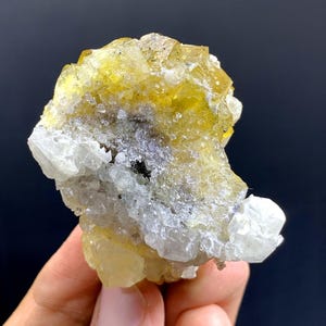 May include: A close-up of a mineral specimen with yellow, translucent crystals. The crystals are mixed with white and grey formations, creating a textured appearance. The mineral is held in a hand against a dark background.