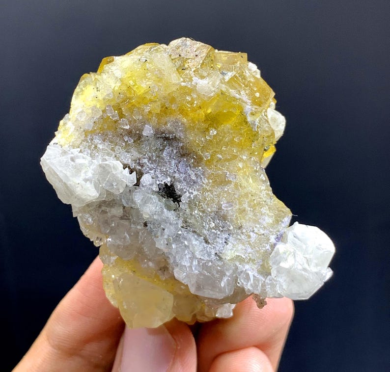 May include: A close-up of a mineral specimen with yellow, translucent crystals. The crystals are mixed with white and grey formations, creating a textured appearance. The mineral is held in a hand against a dark background.