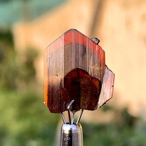Puede incluir: Primer plano de un cristal rojo y marrón con una superficie estriada. El cristal está montado en un soporte de metal.
