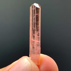 May include: A slender, pink crystal with a faceted top. The crystal is a pale, translucent pink with vertical striations. The crystal is held between two fingers against a dark background.