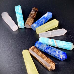 May include: A collection of ten polished crystal wands arranged in a circular pattern. The stones feature various colours including pale pink, bright blue, brown striped, deep royal blue, and translucent yellow, each with a hexagonal point.