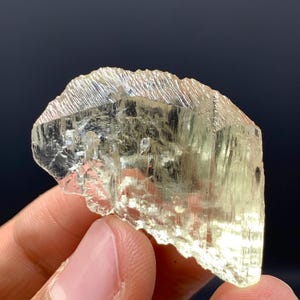 Könnte beinhalten: Ein durchscheinender, hellgelber Kristall mit rauer, strukturierter Oberfläche. Der Kristall hat ein geschichtetes Aussehen mit sichtbaren Streifen. Die Gesamtform ist unregelmäßig, mit gezackten Kanten. Der Kristall wird vor einem dunklen Hintergrund gehalten.