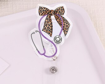 Puede incluir: Un carrete de insignia transparente con un lazo con estampado de leopardo y un diseño de estetoscopio morado. El carrete de la insignia tiene una correa de plástico transparente y un cierre a presión de metal. Una tarjeta blanca dice "My stuff".
