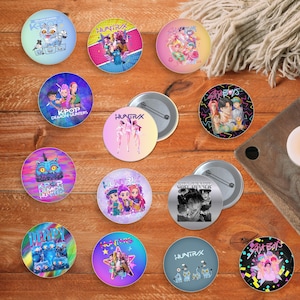 Może przedstawiać: Kolekcja kolorowych, okrągłych przypinek z różnymi wzorami, w tym postaciami z kreskówek, ilustracjami w stylu anime i tekstem. Przypinki są ułożone na drewnianej powierzchni, niektóre z widocznymi tyłami.