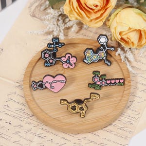 May include: A collection of five enamel pins featuring colorful molecular structures. The pins are arranged on a round wooden tray, with a backdrop of yellow roses and vintage paper. The pins include a pink heart-shaped design and other geometric shapes.
