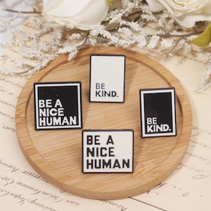 Może przedstawiać: Cztery prostokątne przypinki z czarno-białymi wzorami. Dwie przypinki z napisem "BE A NICE HUMAN" i dwie "BE KIND". Przypinki ułożone są na okrągłej, jasnobrązowej drewnianej tacy, z kwiatowymi akcentami w tle.
