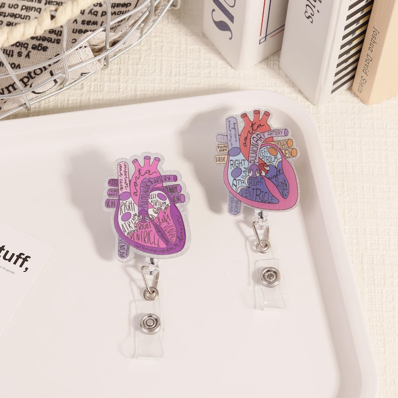 May include: Two clear, heart-shaped badge reels with anatomical diagrams in pink and purple. The diagrams include labels like "Right Atrium" and "Ventricle." Each reel has a retractable cord and a plastic clip.