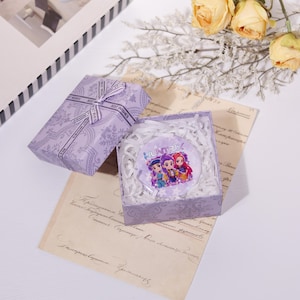 May include: A gift box and a round object with the word "HUNTRIX" and cartoon characters. The gift box is wrapped in lavender paper with a floral pattern and a "Best Wishes" ribbon. The round object is in a clear box with white paper shreds.