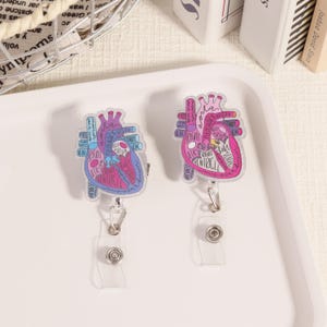 May include: Two heart-shaped badge reels with anatomical diagrams. One is blue and the other is pink, both with labels like "Right Atrium" and "Ventricle." Each reel has a clear plastic strap and a metal clip.