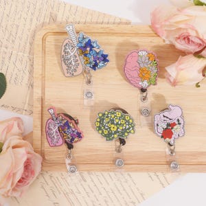 Può includere: Una collezione di porta badge retrattili a tema medico. I disegni includono un polmone, un cervello, uno stomaco e altri organi, ciascuno adornato con accenti floreali o a farfalla colorati. I porta badge sono su un vassoio di legno.