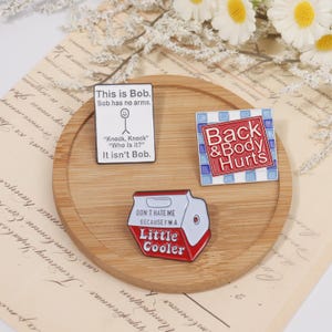May include: Three enamel pins on a wooden tray. One pin reads "This is Bob. Bob has no arms." with a stick figure. Another says "Back & Body Hurts." The third pin is a red and white cooler that says "Little Cooler."