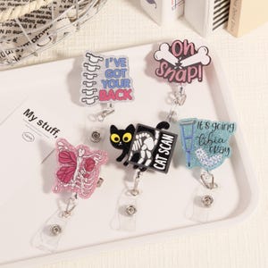 May include: A selection of novelty badge reels with medical and anatomical designs. The reels feature phrases like "I've Got Your Back", a cat with "CAT SCAN", a butterfly with a skeleton, a broken bone with "Oh Snap!", and a leg with "It's going tibia okay".