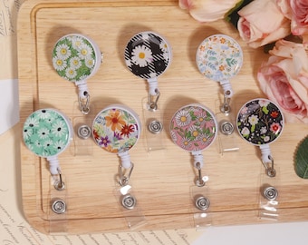 May include: A collection of seven floral badge reels with various designs, including daisies, wildflowers, and abstract patterns. The reels are round with a clear plastic strap and a metal clip. The background includes a wooden tray and floral accents.