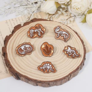 May include: A collection of six dinosaur-themed enamel pins. The pins feature white dinosaur skeletons and a footprint design set against a brown background. The pins are arranged on a wooden slice, with white flowers in the background.