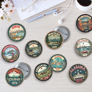 May include: A selection of circular, vintage-style state button badges. Each badge displays a state name and a scenic illustration, including mountains, landmarks, or wildlife. The badges are in various colours and have a metal pin back.