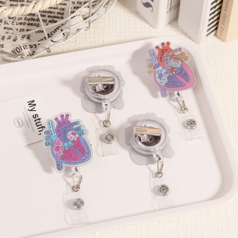 May include: Four retractable badge holders with heart-shaped designs. The hearts are pink, purple, and blue, with anatomical details and text. The badge reels are clear with silver clips and a clear plastic strap. The tray has the text "My stuff" on a card.