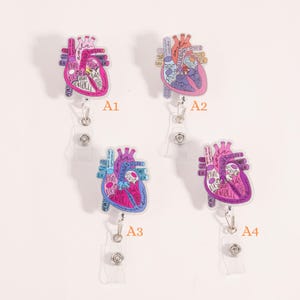 May include: Four anatomical heart-shaped badge reels. Each reel features a detailed illustration of a heart with labeled parts in shades of pink, purple, and blue. The reels have a clear plastic retractable cord and a metal clip.