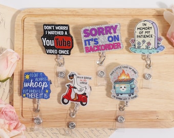 Op de afbeelding: Een verzameling acryl badge reels met humoristische zinnen en afbeeldingen. Ontwerpen omvatten een YouTube-logo, een grafsteen, een gans op een scooter en een vuilnisbak met vlammen. Elke reel heeft een doorzichtige plastic clip.