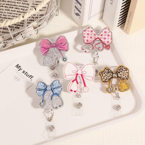 May include: Five badge reels featuring bow and stethoscope designs. The bows are in shades of pink, blue, white, and leopard print. Each reel has a clear plastic clip and a retractable cord, ideal for medical professionals.