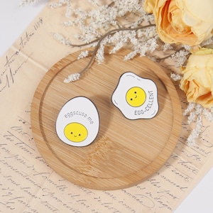 May include: Two enamel pins with egg designs on a wooden tray. One pin says "eggscuse me" and the other says "EGG-CELLENT". Both pins have a white background with a yellow yolk and a smiling face.