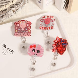 Puede incluir: Una colección de carretes de insignias con temática médica. Los carretes presentan diseños de un útero con el texto "AT YOUR CERVIX", un cerebro con un lazo y un corazón rojo brillante con la palabra "Cardiology".