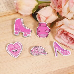 May include: A collection of pink enamel pins featuring a roller skate, boxing glove, heart, leopard print hat, and high heel shoe. The pins have a silver outline and are set against a wooden surface with pink flowers.