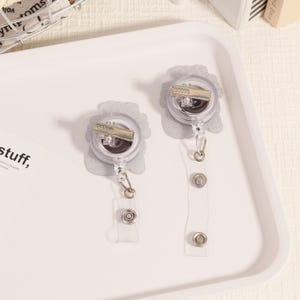 May include: Two clear retractable badge holders with silver accents. The badge reels have a floral design and a metal clip. The clear plastic straps have snap closures. The badge holders are on a white tray.