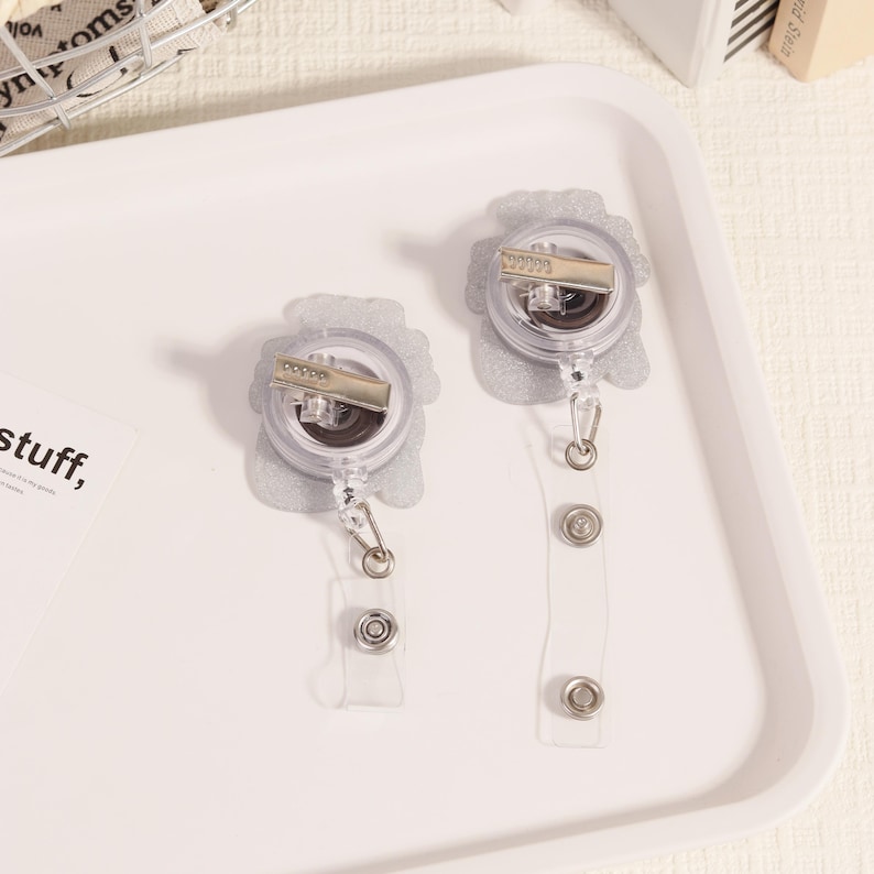 May include: Two clear retractable badge holders with silver accents. The badge reels have a floral design and a metal clip. The clear plastic straps have snap closures. The badge holders are on a white tray.