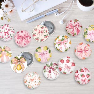 May include: A collection of decorative round pinback buttons with various fruit and bow designs. The buttons feature cherries, lemons, oranges, and strawberries, with pink bows on a white background. Some buttons have a metal backing.