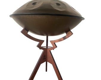 Puede incluir: Un handpan marrón con un borde negro se encuentra sobre un soporte de trípode de madera. El soporte tiene tres patas y una base circular.