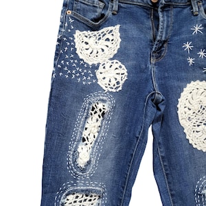 May include: Blue denim jeans with white crochet patches and stitching. The jeans have a distressed look and are decorated with white crochet flowers and stars.