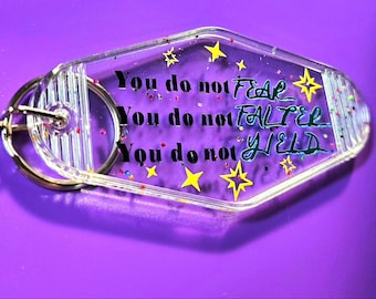 May include: A clear plastic motel key tag with glitter and stars. The text on the tag reads "You do not fear. You do not falter. You do not yield."