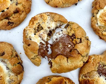 Puede incluir: Primer plano de seis galletas grandes de color marrón dorado con relleno de chocolate derretido y malvavisco tostado. Las galletas están dispuestas sobre una superficie blanca.