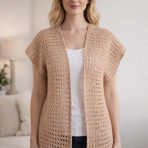 May include: A light brown, open-front crochet cardigan with short sleeves. The cardigan is worn over a white top and paired with blue jeans. The open weave of the cardigan is visible.