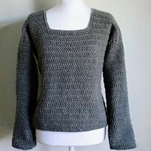 May include: A grey, square-neck crochet sweater. The long-sleeved top is displayed on a white mannequin. The sweater's texture is visible, showcasing the handmade crochet stitches. The colour is a solid, neutral grey.
