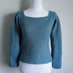 Puede incluir: Un suéter de ganchillo de color turquesa con cuello cuadrado y mangas largas, exhibido en un maniquí blanco. La puntada de ganchillo texturizada es visible. Las mangas tienen puños acanalados.