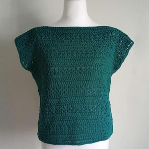May include: A dark teal crochet top with a boat neck and short sleeves. The top is displayed on a white mannequin. The handmade garment has a textured appearance, showcasing the intricate stitch work. The top is a solid color and appears to be made of cotton.