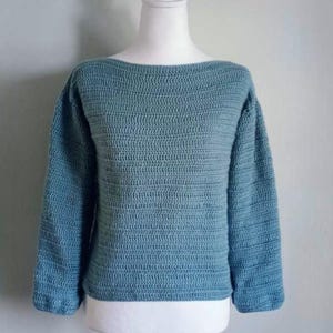 Puede incluir: Un suéter azul verdoso con cuello barco y mangas largas. El suéter se muestra en un maniquí blanco. La textura de punto es visible, con canalé horizontal en el cuerpo y las mangas. Las mangas son de tres cuartos.
