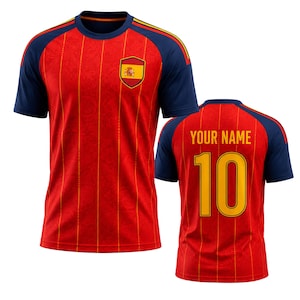 Könnte beinhalten: Rotes und gelbes Fußballtrikot mit marineblauen Ärmeln und Akzenten. Die Vorderseite zeigt das spanische Flaggenemblem und die Rückseite die Nummer 10 und den Text "YOUR NAME" in Gelb.